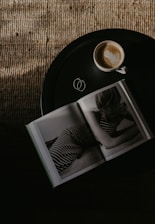 A round black table holds an open book featuring a black-and-white photograph of a person in striped clothing. Beside the book sits a cup of coffee with latte art on top. The table is set against a textured, woven surface that is partially illuminated by light.