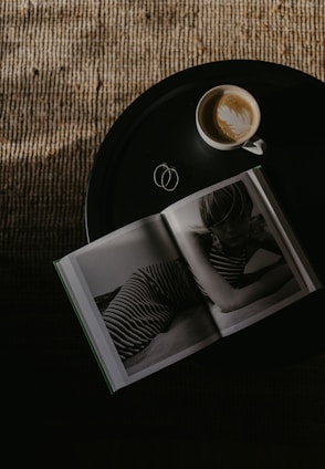 A round black table holds an open book featuring a black-and-white photograph of a person in striped clothing. Beside the book sits a cup of coffee with latte art on top. The table is set against a textured, woven surface that is partially illuminated by light.