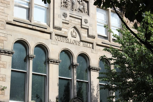 A historical building facade made of stone, featuring arched windows and decorative carvings. The engraving 'Oddfellows Hall' is prominently displayed above the windows. Surrounding the building are lush green trees partially obscuring the view.