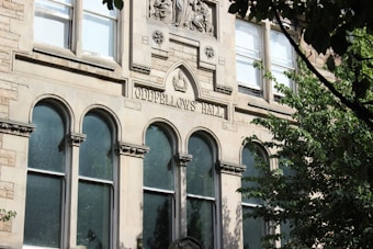 A historical building facade made of stone, featuring arched windows and decorative carvings. The engraving 'Oddfellows Hall' is prominently displayed above the windows. Surrounding the building are lush green trees partially obscuring the view.