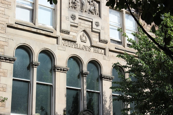 A historical building facade made of stone, featuring arched windows and decorative carvings. The engraving 'Oddfellows Hall' is prominently displayed above the windows. Surrounding the building are lush green trees partially obscuring the view.