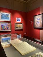 A gallery space featuring several framed paintings on a bold red wall, each showcasing vibrant colors and distinct artistic styles. A display case beneath the paintings holds an open sketchbook or large book, partially visible.