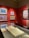 A gallery space featuring several framed paintings on a bold red wall, each showcasing vibrant colors and distinct artistic styles. A display case beneath the paintings holds an open sketchbook or large book, partially visible.