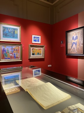 A gallery space featuring several framed paintings on a bold red wall, each showcasing vibrant colors and distinct artistic styles. A display case beneath the paintings holds an open sketchbook or large book, partially visible.