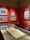 A gallery space featuring several framed paintings on a bold red wall, each showcasing vibrant colors and distinct artistic styles. A display case beneath the paintings holds an open sketchbook or large book, partially visible.