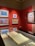 A gallery space featuring several framed paintings on a bold red wall, each showcasing vibrant colors and distinct artistic styles. A display case beneath the paintings holds an open sketchbook or large book, partially visible.