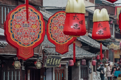 Red Chinese lanterns and decorative ornaments hang in a traditional street, featuring intricate patterns and calligraphy. The background includes historic architecture with tiled roofs and wooden structures.