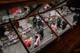 A lively outdoor market scene viewed from above, with colorful stalls selling various goods such as toys, snacks, and souvenirs. People, including children and adults, are browsing and shopping, dressed warmly in coats. The scene is vibrant with activity and chatter, showcasing a bustling marketplace atmosphere.