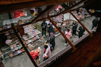 A lively outdoor market scene viewed from above, with colorful stalls selling various goods such as toys, snacks, and souvenirs. People, including children and adults, are browsing and shopping, dressed warmly in coats. The scene is vibrant with activity and chatter, showcasing a bustling marketplace atmosphere.