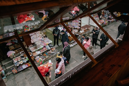 A lively outdoor market scene viewed from above, with colorful stalls selling various goods such as toys, snacks, and souvenirs. People, including children and adults, are browsing and shopping, dressed warmly in coats. The scene is vibrant with activity and chatter, showcasing a bustling marketplace atmosphere.