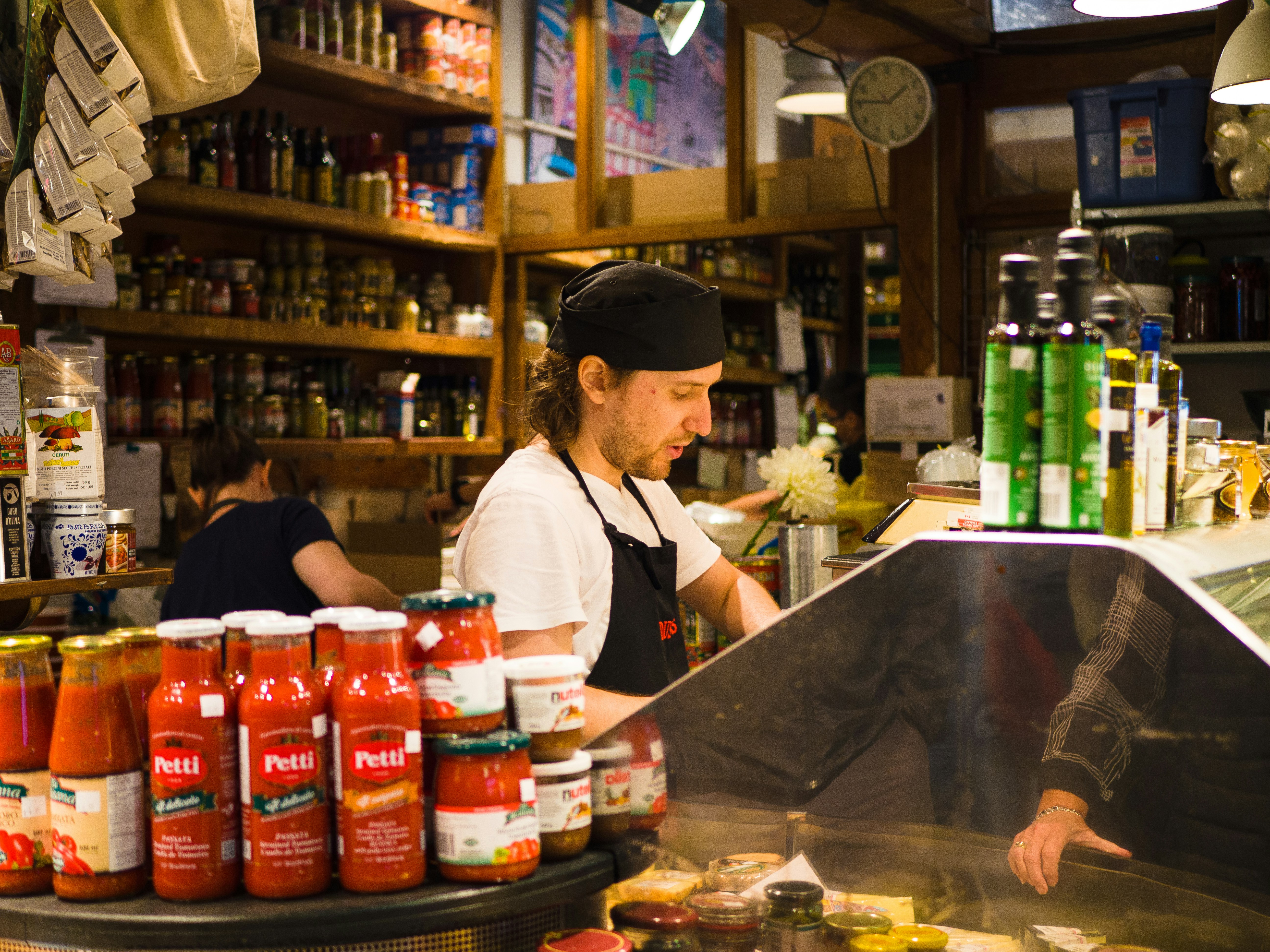 A grocery category manager staring at a cluttered Excel spreadsheet on a monitor, surrounded by paper inventory sheets, while a colleague in the background reviews an AI dashboard on a tablet showing green availability metrics