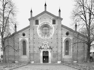 A historic brick church with a large circular stained-glass window at the center, flanked by two tall arched windows. The entrance is adorned with intricate stone carvings. Leafless trees frame the building, and the foreground is a cobblestone path.