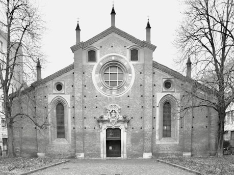 A historic brick church with a large circular stained-glass window at the center, flanked by two tall arched windows. The entrance is adorned with intricate stone carvings. Leafless trees frame the building, and the foreground is a cobblestone path.