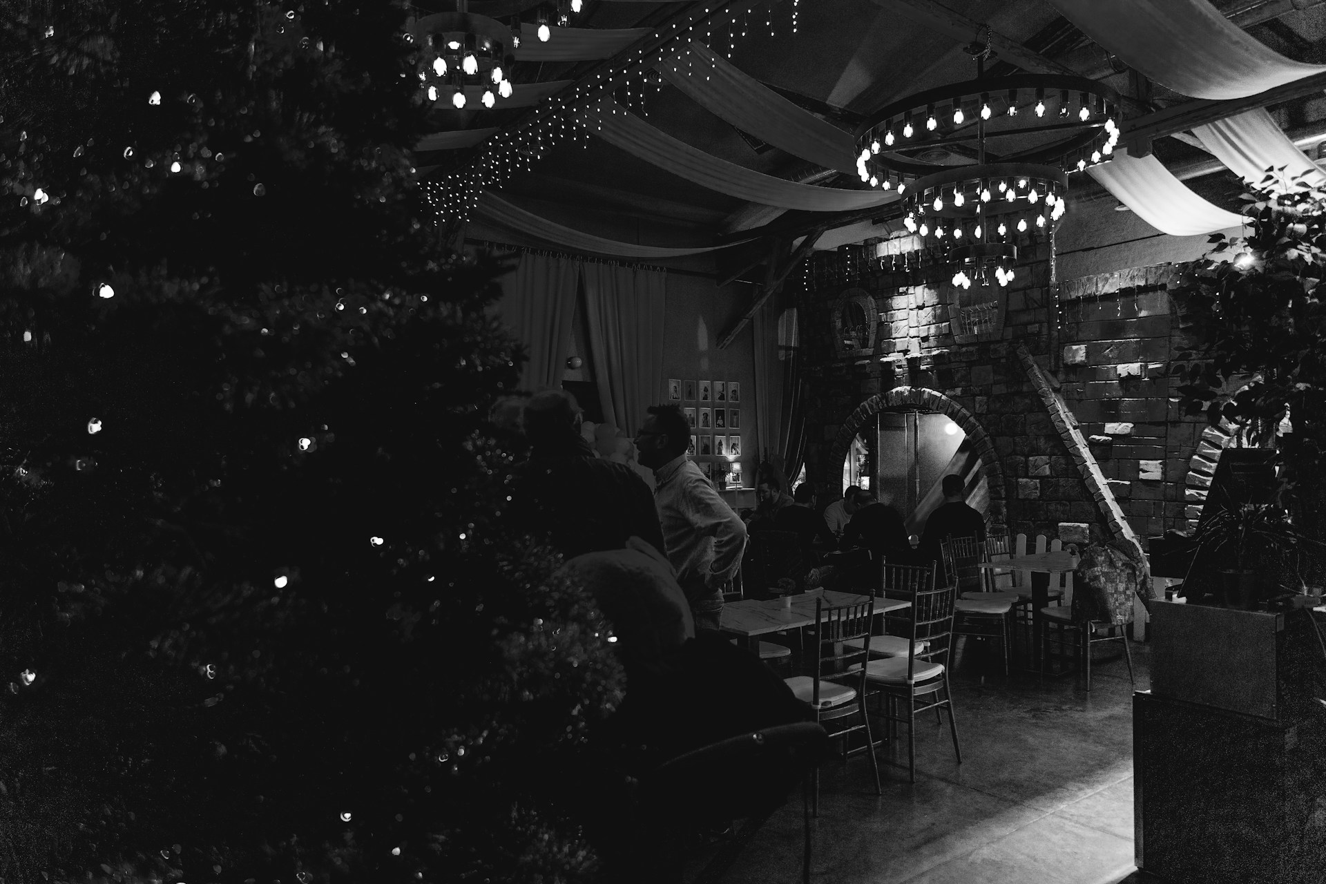 A dimly lit indoor space with a cozy and rustic atmosphere. The room features a large, decorated Christmas tree and ambient lighting provided by strings of small lights and a circular chandelier. Stone walls and an archway contribute to the old-world charm. Several people are gathered around tables, adding to the festive and social setting.