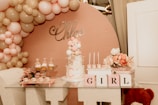 A decorative setup featuring a pastel-colored balloon arch with pink, beige, and white balloons. A cake adorned with small teddy bear figures and pastel balloons sits on a glass table. Nearby, cupcakes are arranged on a tiered tray, and flowers in shades of pink and white provide additional decoration. Blocks spelling out 'GIRL' and a teddy bear are included in the arrangement.