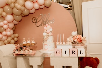 A decorative setup featuring a pastel-colored balloon arch with pink, beige, and white balloons. A cake adorned with small teddy bear figures and pastel balloons sits on a glass table. Nearby, cupcakes are arranged on a tiered tray, and flowers in shades of pink and white provide additional decoration. Blocks spelling out 'GIRL' and a teddy bear are included in the arrangement.