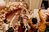 A lively celebration featuring a group of children reaching towards bubbles, surrounded by a beautiful arrangement of pink and beige balloons. An adult holds a child in their arms in the center, possibly guiding her to interact with the bubbles. The background includes decorative elements and possibly a sign.