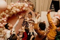 A lively celebration featuring a group of children reaching towards bubbles, surrounded by a beautiful arrangement of pink and beige balloons. An adult holds a child in their arms in the center, possibly guiding her to interact with the bubbles. The background includes decorative elements and possibly a sign.