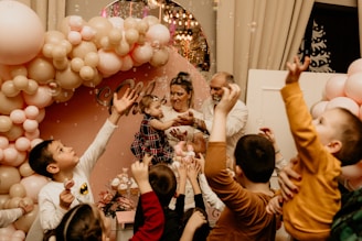 A lively celebration featuring a group of children reaching towards bubbles, surrounded by a beautiful arrangement of pink and beige balloons. An adult holds a child in their arms in the center, possibly guiding her to interact with the bubbles. The background includes decorative elements and possibly a sign.