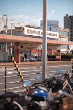 A street scene featuring a tram station with signs in Japanese. Several bicycles are parked in the foreground, and people can be seen sitting on benches near the station. Buildings and electrical wires are visible in the background, indicating an urban setting.
