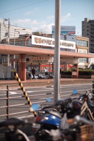 A street scene featuring a tram station with signs in Japanese. Several bicycles are parked in the foreground, and people can be seen sitting on benches near the station. Buildings and electrical wires are visible in the background, indicating an urban setting.