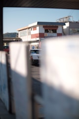 A partially obscured view of a street scene featuring a white taxi with a blue sign on top. The background displays a building with red and white exterior and a signage with characters, suggesting a possible commercial area. The foreground is blurred, focusing on the central taxi.