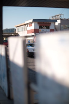 A partially obscured view of a street scene featuring a white taxi with a blue sign on top. The background displays a building with red and white exterior and a signage with characters, suggesting a possible commercial area. The foreground is blurred, focusing on the central taxi.
