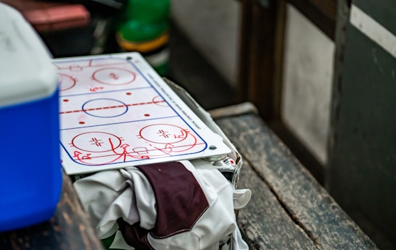 A coaching board displaying a diagram of a hockey rink is placed on a wooden bench. There is a blue cooler to the left and a piece of clothing, possibly a jersey, draped over the bench. A green object is visible in the background, slightly out of focus.