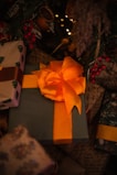A large gift wrapped in green paper and adorned with a striking orange bow is situated among other presents. Surrounding the gifts are festive decorations, including red berries and greenery, under the warm glow of soft, ambient lights.