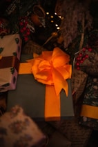 A large gift wrapped in green paper and adorned with a striking orange bow is situated among other presents. Surrounding the gifts are festive decorations, including red berries and greenery, under the warm glow of soft, ambient lights.