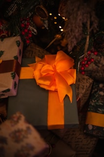 A large gift wrapped in green paper and adorned with a striking orange bow is situated among other presents. Surrounding the gifts are festive decorations, including red berries and greenery, under the warm glow of soft, ambient lights.