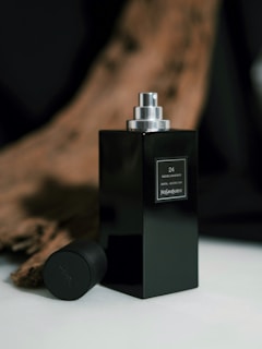 A sleek black perfume bottle with a metallic spray top is positioned in the foreground, accompanied by its matching cap lying nearby. The background features an artistic blur of warm, wooden tones, adding a sophisticated touch to the composition.