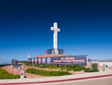 A large white cross stands prominently on a platform surrounded by a memorial. The site includes a sign reading 'Mt. Soledad National Veterans Memorial' with various military emblems displayed. There are manicured bushes and an American flag. The sky is clear and blue, enhancing the solemn and respectful ambiance.