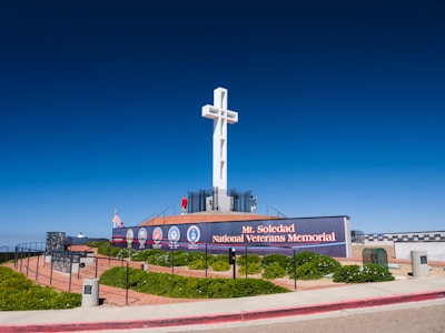 A large white cross stands prominently on a platform surrounded by a memorial. The site includes a sign reading 'Mt. Soledad National Veterans Memorial' with various military emblems displayed. There are manicured bushes and an American flag. The sky is clear and blue, enhancing the solemn and respectful ambiance.
