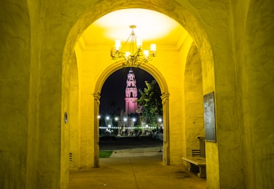 A warmly lit, arched hallway leads to an outdoor scene featuring a historical tower illuminated at night. Soft yellow light fills the corridor, highlighting textured walls and a decorative chandelier. The tower, visible through the archway, is adorned with pink lighting and surrounded by trees and white lights.