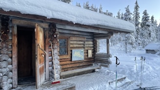A wooden log cabin stands in a snowy landscape, with a partially open door and a snow-covered roof. The surrounding area is filled with snow-laden trees. Ski poles are sticking out of the snow next to the cabin, and a map is mounted on the cabin wall.