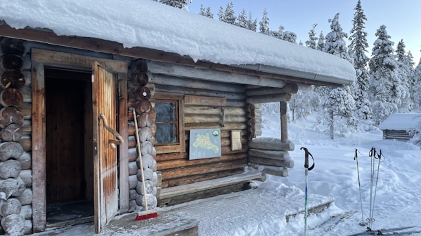 A wooden log cabin stands in a snowy landscape, with a partially open door and a snow-covered roof. The surrounding area is filled with snow-laden trees. Ski poles are sticking out of the snow next to the cabin, and a map is mounted on the cabin wall.