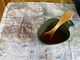 A detailed topographic map is laid out on a wooden table. On top of the map, there is a green camping mug filled with a brown liquid, and an orange plastic spork is resting inside the mug.