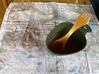 A detailed topographic map is laid out on a wooden table. On top of the map, there is a green camping mug filled with a brown liquid, and an orange plastic spork is resting inside the mug.