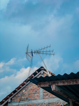 An outdoor scene featuring a rooftop with a TV antenna mounted on it. The rooftop is made of bricks with a tiled edge, and the antenna is angled against a blue sky with light clouds.