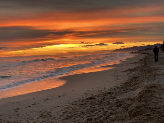 A sandy beach at sunset with the ocean waves gently rolling in. The sky is painted with vibrant shades of orange, yellow, and red as the sun descends towards the horizon. Silhouettes of several people are visible along the shoreline, capturing the serene moment.