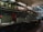 A dimly lit interior of a market or shop area with closed metal shutters. Above the shutters, there are signs with Chinese characters. The space appears empty, with some items hanging and scattered on the counters.