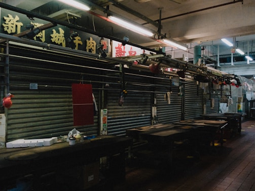 A dimly lit interior of a market or shop area with closed metal shutters. Above the shutters, there are signs with Chinese characters. The space appears empty, with some items hanging and scattered on the counters.