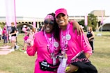 Two people are smiling and posing together outdoors, wearing matching bright pink shirts and accessories, possibly participating in a charity event or walk. Both individuals appear happy and relaxed, and there are other participants and banners in the background, suggesting a lively and supportive atmosphere.
