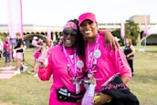 Two people are smiling and posing together outdoors, wearing matching bright pink shirts and accessories, possibly participating in a charity event or walk. Both individuals appear happy and relaxed, and there are other participants and banners in the background, suggesting a lively and supportive atmosphere.