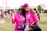 Two people are smiling and posing together outdoors, wearing matching bright pink shirts and accessories, possibly participating in a charity event or walk. Both individuals appear happy and relaxed, and there are other participants and banners in the background, suggesting a lively and supportive atmosphere.