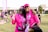 Two people are smiling and posing together outdoors, wearing matching bright pink shirts and accessories, possibly participating in a charity event or walk. Both individuals appear happy and relaxed, and there are other participants and banners in the background, suggesting a lively and supportive atmosphere.