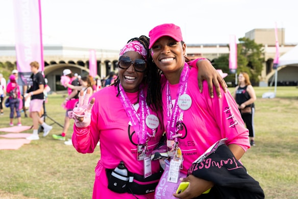 Two people are smiling and posing together outdoors, wearing matching bright pink shirts and accessories, possibly participating in a charity event or walk. Both individuals appear happy and relaxed, and there are other participants and banners in the background, suggesting a lively and supportive atmosphere.