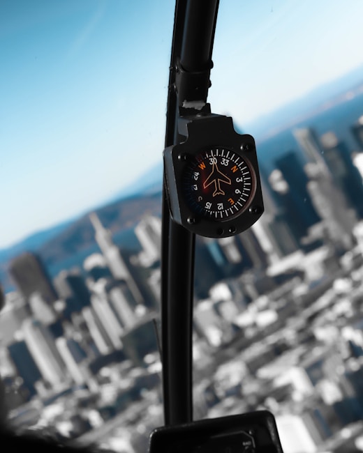 An aircraft compass is prominently displayed with a blurred cityscape visible through the window, indicating a scene from inside a cockpit. The compass is mounted on a black panel, showing cardinal directions and degree markings.