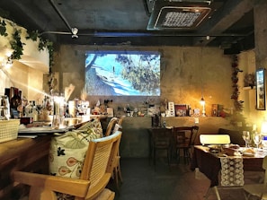 A cozy cafe or restaurant interior with wooden chairs and tables, decorated with various ornaments and books on shelves. Soft lighting provides a warm atmosphere, enhanced by a video projection on the wall showing a scenic outdoor image.
